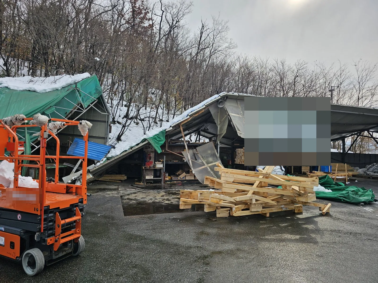 눈에 무너진 옥외 가설 구조물 앞에 고소작업차와 목재 잔재가 있다.