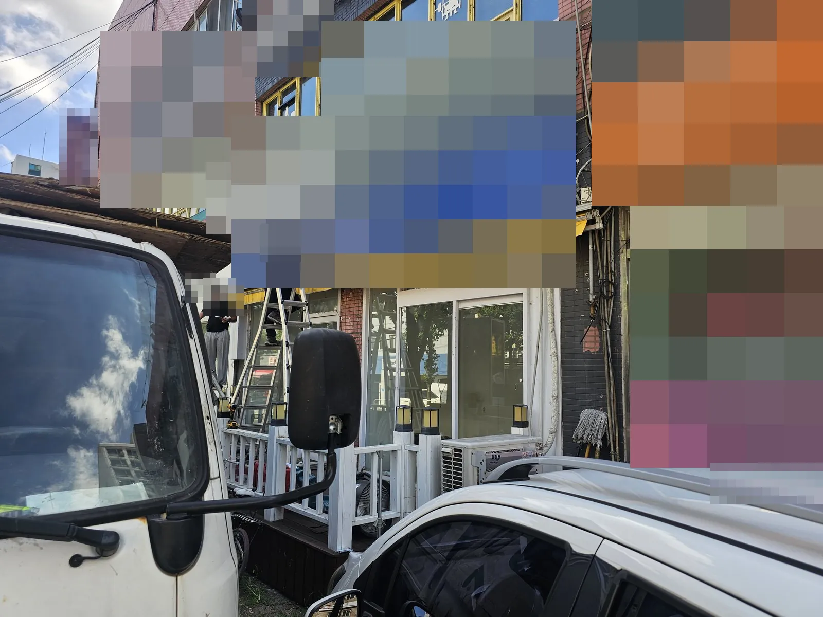 전경에 차량들이 있고 중앙에 사다리가 보인다. 사람의 몸 일부가 보이며 머리·얼굴 부근에는 모자이크가 겹쳐 있다. 유리 출입문·실외기·낮은 울타리 형태가 보인다. 상단과 오른쪽의 넓은 영역이 모자이크로 가려져 있다. 유리와 차량 창에 주변 구조물 등의 반사가 보인다.