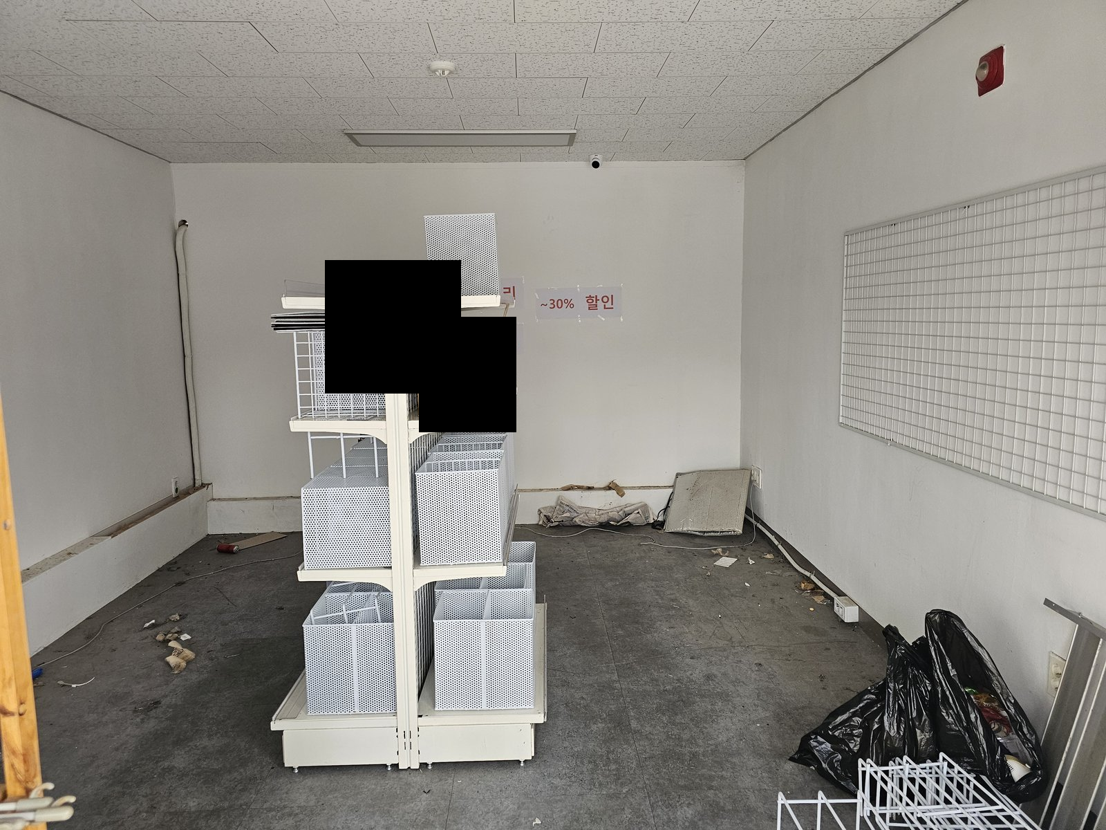 비어 있는 매장 실내에 흰색 진열대와 바구니, 쓰레기 봉투가 있다. 벽에 할인 문구 종이가 붙어 있고 일부는 검은 가림 처리되어 있다. 사진에 보이지 않는 범위나 과거 작업 이력은 확인하지 않았습니다.