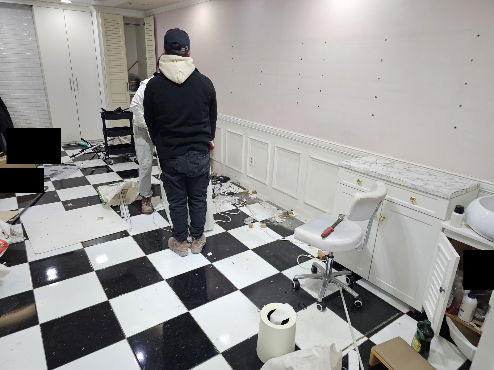 체스판 무늬 바닥의 실내에서 철거 작업 중인 사람들이 뒷모습으로 보인다. 흰색 수납장, 의자, 잔해가 보인다. 사진에 보이지 않는 범위나 과거 작업 이력은 확인하지 않았습니다.