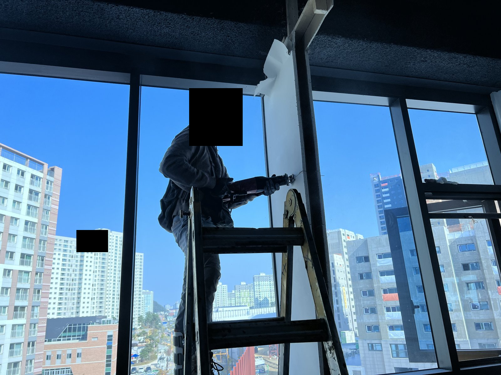 실내 유리창 앞 사다리 위에서 작업자가 전동공구로 작업한다. 얼굴은 검은 가림으로 덮여 있고 창밖에 건물이 보인다. 사진에 보이지 않는 범위나 과거 작업 이력은 확인하지 않았습니다.